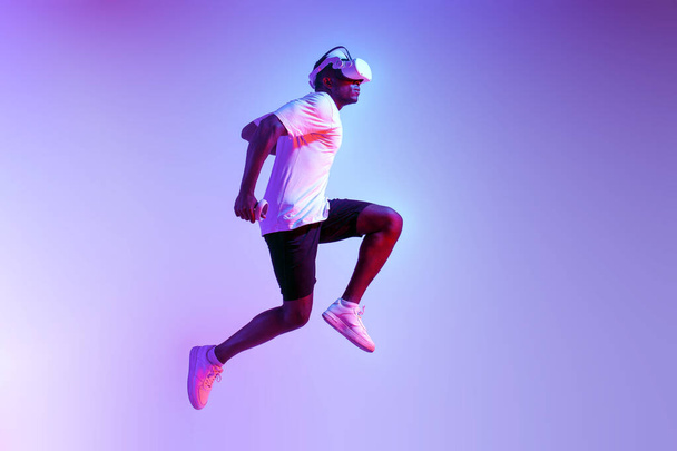 hombre atlético afroamericano corre rápidamente y salta en gafas de realidad virtual en iluminación de neón, el atleta chico entrena y se mueve en gafas vr y sostiene joysticks - Foto, imagen