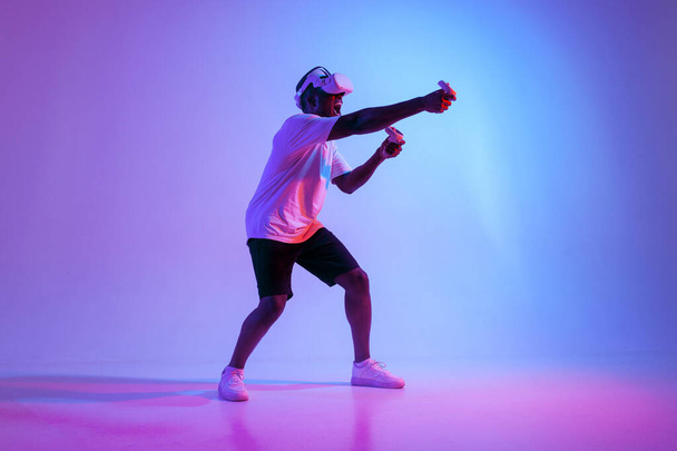 african american man in virtual reality glasses aiming and shooting in neon lighting, guy gamer playing and fighting in vr glasses with joysticks - Photo, Image