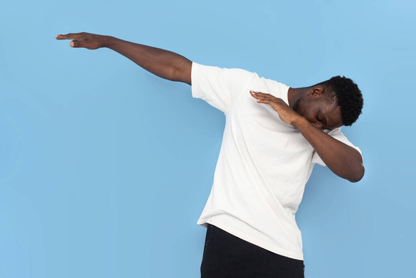 jong Afrikaans amerikaans man in wit t-shirt danst en maakt dab beweging op blauw geïsoleerde achtergrond, man bedekt zijn gezicht met zijn handen en wijst naar de zijkant - Foto, afbeelding
