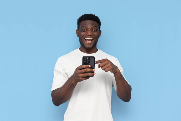 afro americano cara no branco t-shirt usa smartphone e escolhe no azul isolado fundo, homem detém telefone e pressiona o dedo - Foto, Imagem