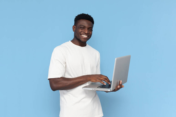 Afrikkalainen amerikkalainen kaveri valkoisessa t-paidassa käyttää kannettavaa tietokonetta ja hymyilee sinisellä taustalla, mies iloitsee ja tyypit tietokoneella - Valokuva, kuva
