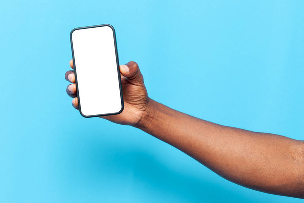main de l'homme afro-américain tenant smartphone avec écran blanc sur fond bleu isolé, la main annonce l'espace de copie sur le téléphone - Photo, image
