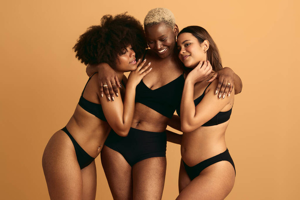Young smiling multiethnic ladies in black lingerie hugging each other in studio against orange background support and acceptance concept - Valokuva, kuva