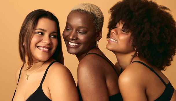 Side view of cheerful young multiracial female models with short and long hair in black underwear, smiling and looking at each other against beige background - Photo, Image