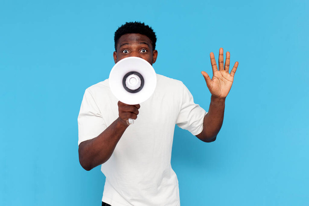 Afrikanisch-amerikanischer Mann im weißen T-Shirt verkündet Informationen in Lautsprecher auf blauem Hintergrund, Mann schreit in Megafon - Foto, Bild