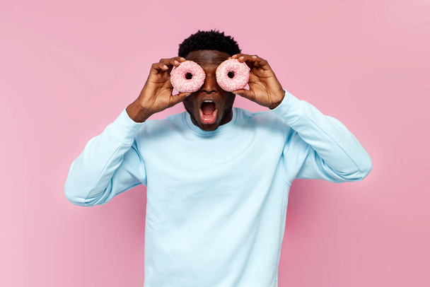 shocked african american man in blue sweater holding sweet donuts on pink isolated background, man with pastries and food screaming and being surprised - Fotoğraf, Görsel