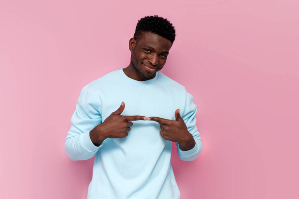modest african american man in blue sweater is shy and smiles on pink isolated background, embarrassed and pleased man - Φωτογραφία, εικόνα