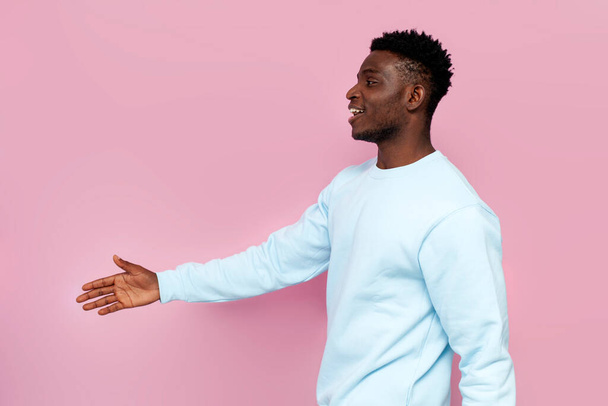 african american man in blue sweater smiles and holds out his hand for handshake on pink isolated background, side view - Foto, immagini
