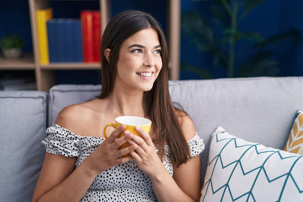 Jeune belle femme hispanique buvant du café assis sur le canapé à la maison - Photo, image