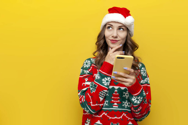 Ragazza pensierosa in maglione di Natale e cappello di Babbo Natale utilizza smartphone e sogni su sfondo giallo isolato, donna tiene il telefono e pensa in abiti di Natale - Foto, immagini