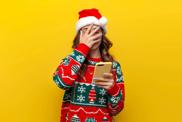 μπερδεμένο κορίτσι με χριστουγεννιάτικο πουλόβερ και καπέλο santa claus χρησιμοποιεί smartphone και δείχνει facepalm σε κίτρινο απομονωμένο φόντο, γυναίκα κρατά τηλέφωνο καλύπτει το πρόσωπό της με το χέρι της στα ρούχα της Πρωτοχρονιάς - Φωτογραφία, εικόνα