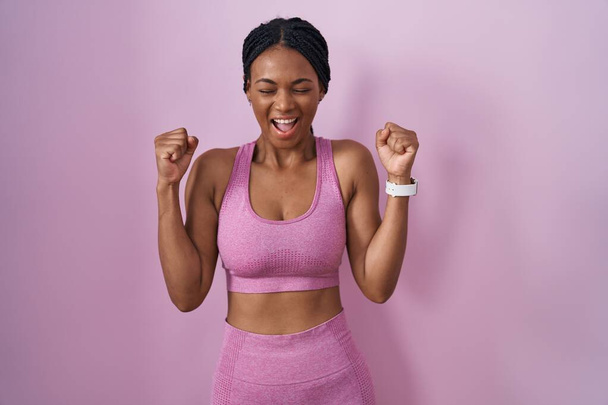Afrikai amerikai nő copfban sportruházatot visel rózsaszín háttér izgatott a siker karját, és csukott szemmel ünnepli győzelem mosolyogva. győztes koncepció.  - Fotó, kép