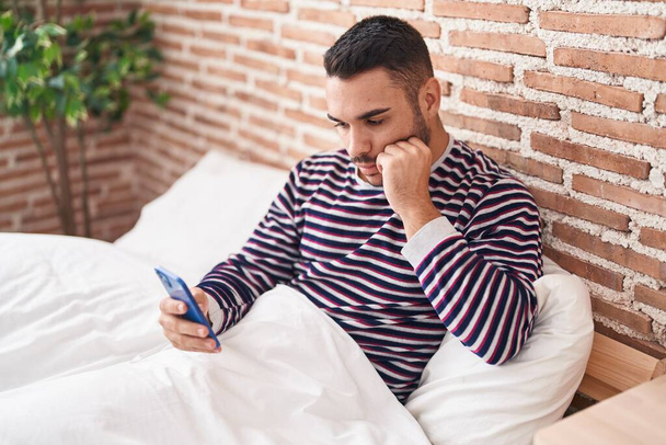 Young hispanic man using smartphone sitting on bed at bedroom - Foto, Imagem