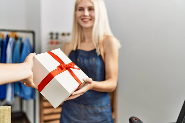 Jonge blonde vrouw winkel assistent glimlachen zelfverzekerd geven geschenk aan de klant in kleding winkel - Foto, afbeelding