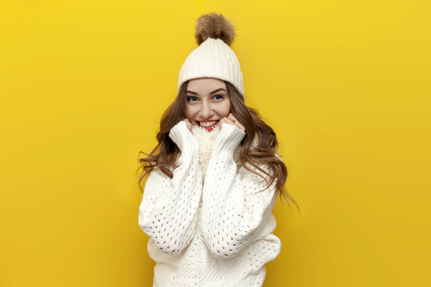 young cute girl in warm soft winter clothes smiles on yellow isolated background, woman in white hat and scarf touches comfortable and cozy knitted sweater - Photo, Image