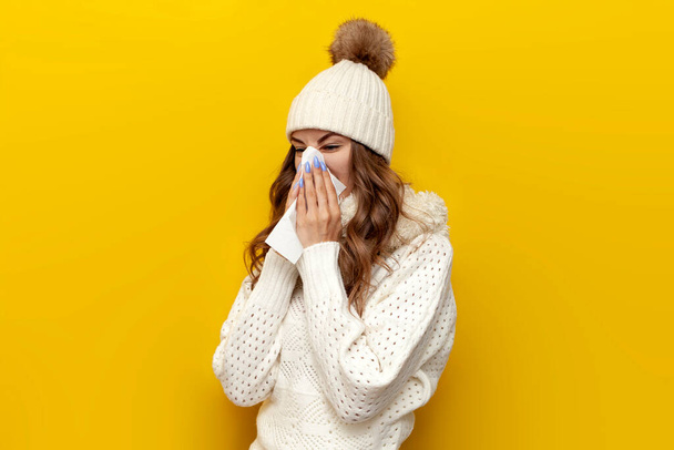 sick girl in warm soft winter clothes with runny nose in napkin on yellow isolated background, cold woman in white hat and scarf in comfortable and cozy knitted sweater shows flu and cold symptoms - Photo, Image