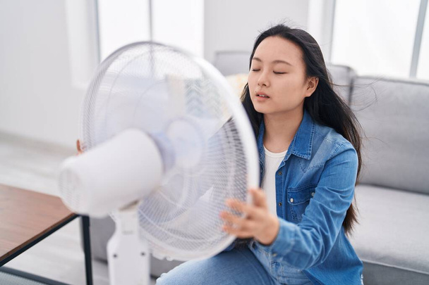 Jonge Chinese vrouw met behulp van ventilator zitten op de bank thuis - Foto, afbeelding