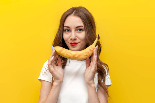 mladá dívka ukazuje políbené banány a naráží na intimitu na žlutém izolovaném pozadí, žena s ovocnými úsměvy, sexy a erotické koncept - Fotografie, Obrázek