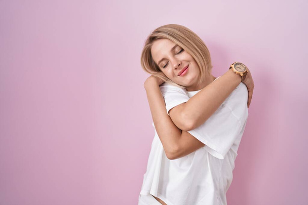 Joven mujer caucásica de pie sobre fondo rosa abrazándose feliz y positivo, sonriendo confiado. amor propio y cuidado personal  - Foto, imagen