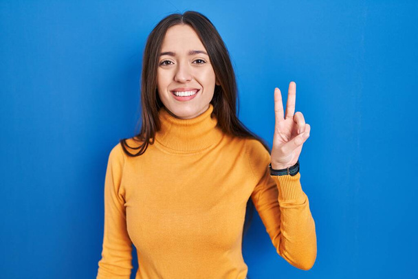 Młoda brunetka kobieta stoi nad niebieskim tle pokazując i wskazując palcem numer dwa uśmiechając się pewny siebie i szczęśliwy.  - Zdjęcie, obraz