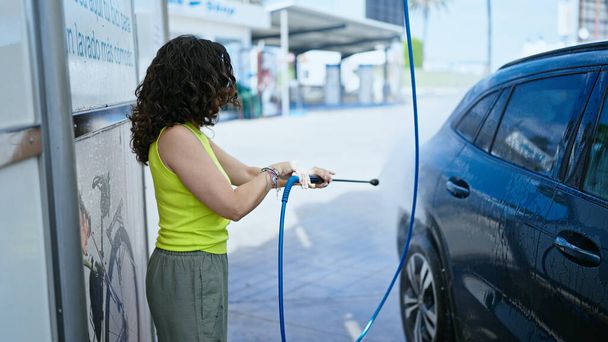 Middle age hispanic woman washing car with pressure washer at car wash station - Photo, Image
