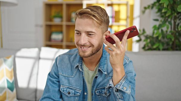 Young caucasian man listening voice message by smartphone sitting on sofa at home - Photo, Image