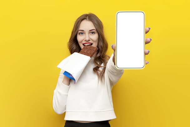 若い女の子は大きいチョコレート バーを食べ,黄色い隔離された背景の空白のスマートフォン スクリーン,甘い食べ物の広告の電話表示が付いている女性を示します - 写真・画像