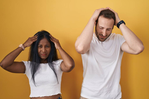 Ein gemischtrassiges Paar, das vor gelbem Hintergrund steht und unter Kopfschmerzen leidet, verzweifelt und gestresst wegen Schmerzen und Migräne. Hände auf den Kopf.  - Foto, Bild