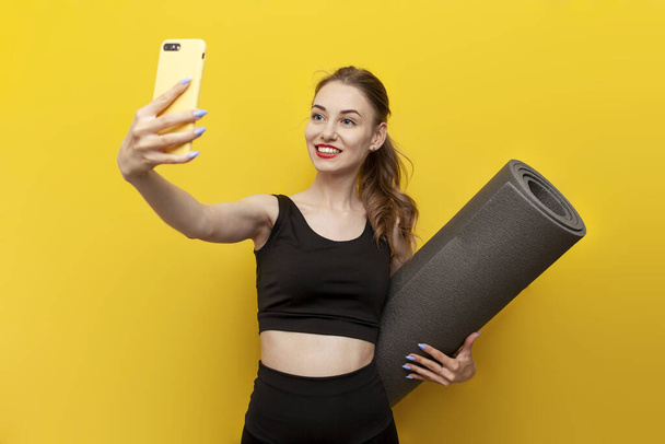 young sports girl in sportswear with yoga mat uses smartphone on yellow isolated background, slender woman takes selfie and communicates by video call - Фото, изображение