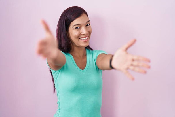 Mujer hispana joven de pie sobre fondo rosa mirando a la cámara sonriendo con los brazos abiertos para abrazarse. expresión alegre abrazando la felicidad.  - Foto, imagen