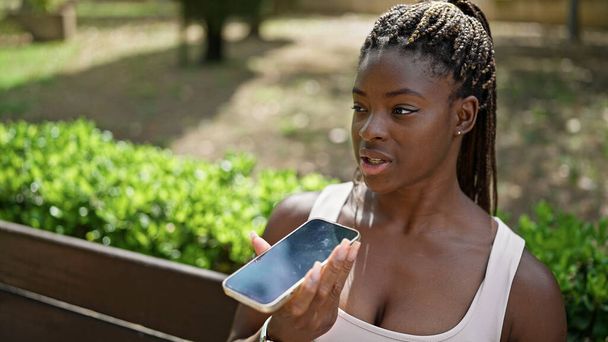 African american woman sending voice message with smartphone sitting on bench at park - Photo, Image