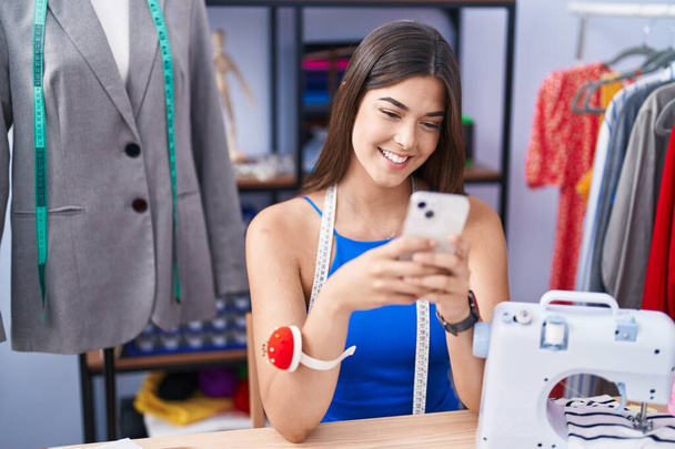 Młoda piękna Latynoska kobieta krawiec uśmiechnięta pewnie za pomocą smartfona w sklepie krawieckim - Zdjęcie, obraz