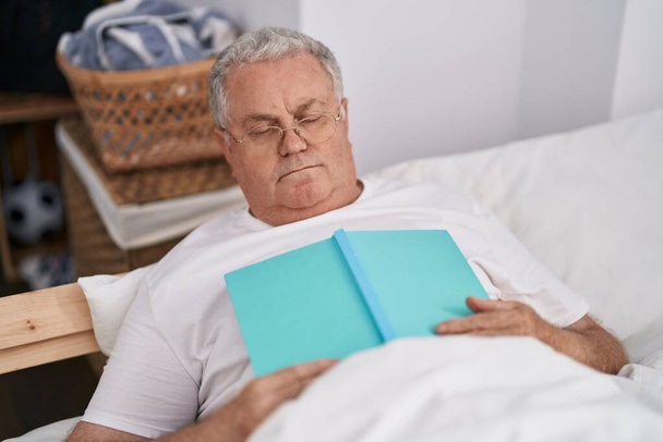 Grauhaariger Mann mittleren Alters liegt schlafend auf Bett und hält Buch im Schlafzimmer - Foto, Bild