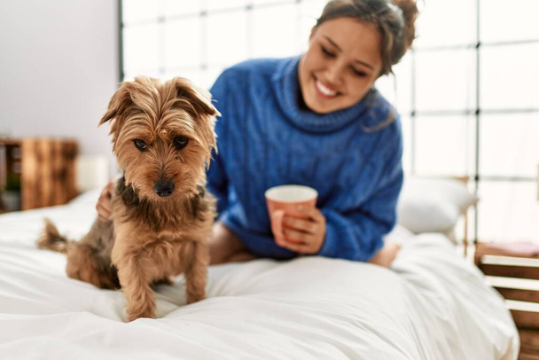 Joven mujer hispana hermosa bebiendo taza de café sentado en la cama con perro en el dormitorio - Foto, imagen