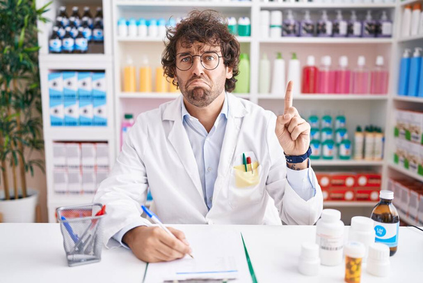 Latino nuori mies työskentelee apteekissa apteekki apteekki osoittaa ylös etsivät surullinen ja järkyttynyt, osoittaa suuntaan sormin, onneton ja masentunut.  - Valokuva, kuva