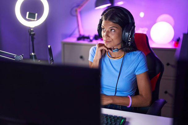 Брюнетка молодая женщина играет в видеоигры серьезное лицо думая о вопросе с рукой на подбородке, вдумчивый о запутанной идее  - Фото, изображение