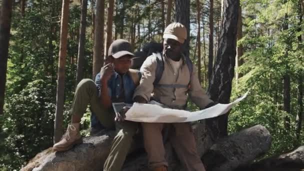 Afrikai-amerikai idős férfi és unokája hátizsákot visel ül a sziklán a hegyi erdőben a nyári nap olvasás térkép - Felvétel, videó