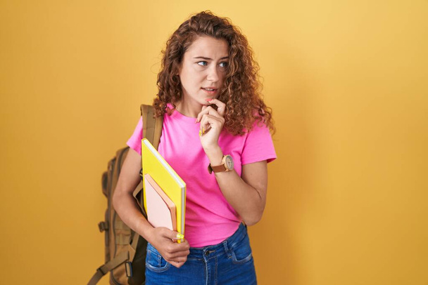 Mujer joven caucásica vistiendo mochila de estudiante y sosteniendo libros pensando preocupado por una pregunta, preocupado y nervioso con la mano en la barbilla  - Foto, Imagen