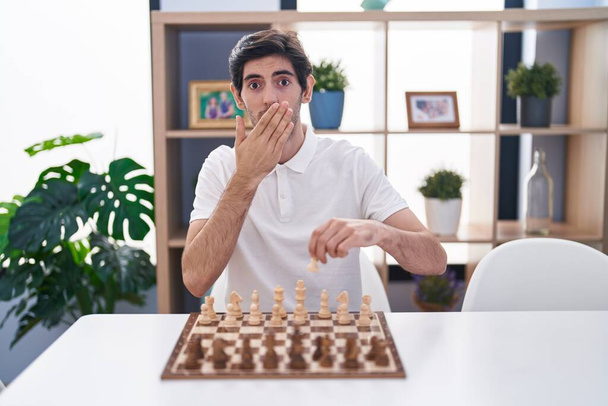 Joven hispano jugando al ajedrez sentado en la mesa cubriendo la boca con la mano, sorprendido y temeroso de equivocarse. expresión sorprendida  - Foto, imagen