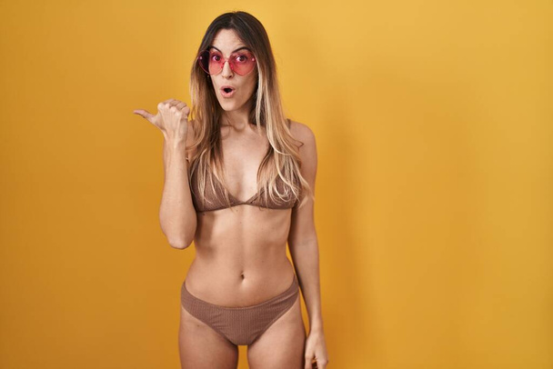 Giovane donna ispanica che indossa bikini su sfondo giallo sorpreso indicando con il dito della mano di lato, bocca aperta espressione stupita.  - Foto, immagini