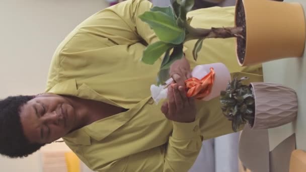 Yetişkin Afro-Amerikan kadının oturma odasındaki çiçekleriyle ilgilenirken su püskürtücü kullandığı dikey bir fotoğraf. - Video, Çekim
