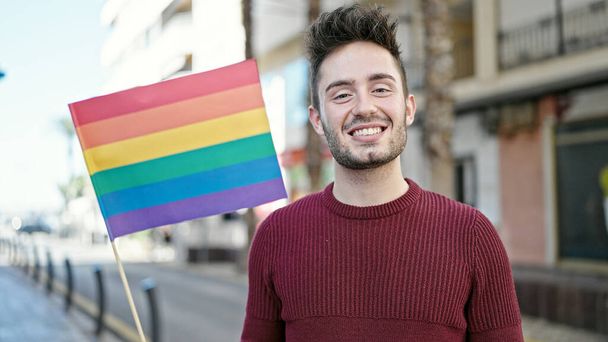 Giovane uomo ispanico sorridente fiducioso tenendo bandiera arcobaleno in strada - Foto, immagini