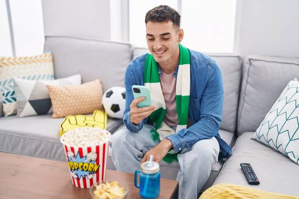 Mladý hispánec muž fotbalový fanoušek pomocí smartphone sedí na pohovce doma - Fotografie, Obrázek