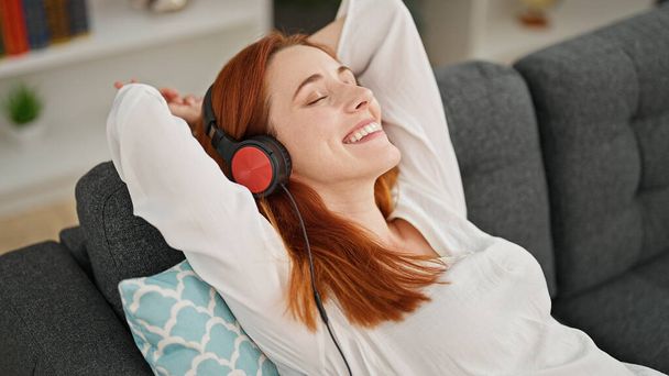 Jeune rousse femme écouter de la musique assis sur le canapé à la maison - Photo, image