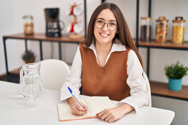 Νέα όμορφη ισπανόφωνη γυναίκα που γράφει στο σημειωματάριο κάθεται στο τραπέζι στο σπίτι - Φωτογραφία, εικόνα