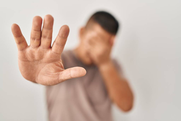 Jeune homme hispanique debout sur fond blanc couvrant les yeux des mains et faisant arrêter geste avec expression triste et peur. concept embarrassé et négatif.  - Photo, image