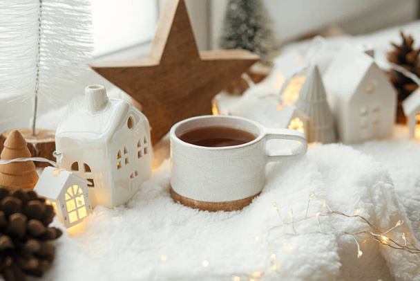 Stijlvol kopje thee met moderne kersthuizen, dennenappel, houten ster- en boomdecor, gouden lampjes op warme deken op vensterbank. Gezellige kerst, Scandinavische stijl. Kerst achtergrond - Foto, afbeelding