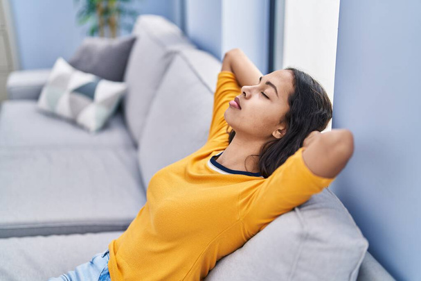 Молода афроамериканська жінка розслаблена руками на голові сидячи вдома на дивані. - Фото, зображення