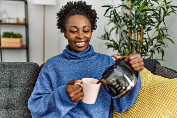 Африканська американка поливає каву на чашку, сидячи вдома на дивані. - Фото, зображення