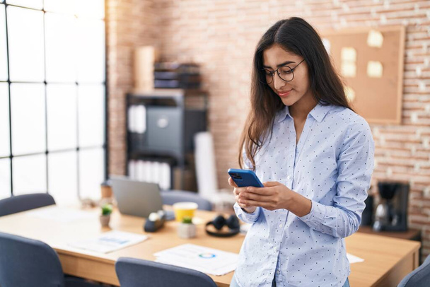 Молода іспаномовна дівчина бізнесмен посміхається впевнено використовуючи смартфон в офісі
 - Фото, зображення
